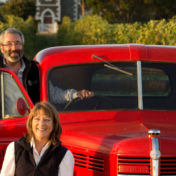 Stephen and Prue Henschke sat next to a red car in the middle of a vinyard