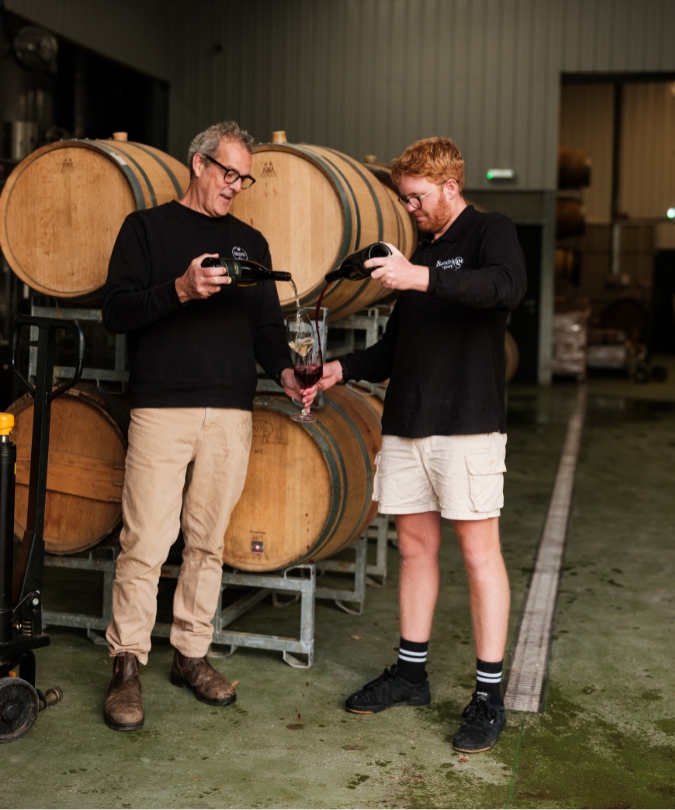 Two men in front of wine barrels pouring wine into their glasses