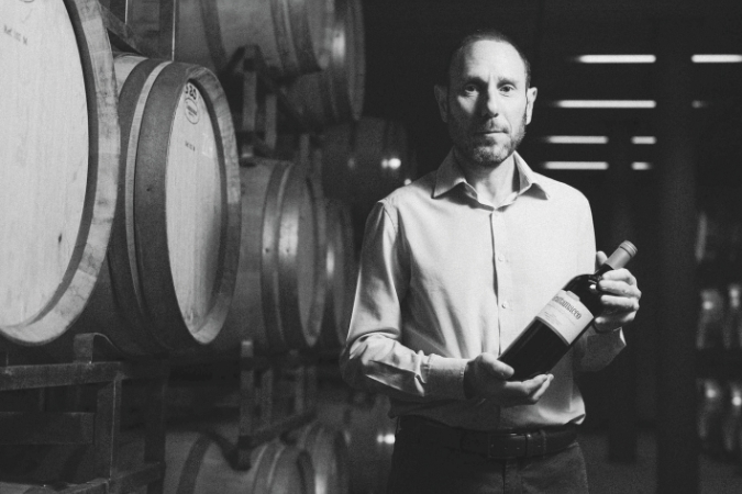Luca Marrone holding a bottle of Grattamacco next to barrels in winery
